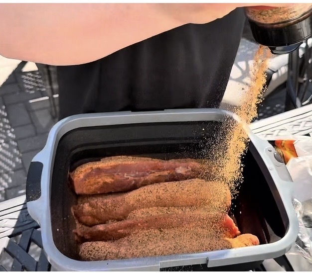 A person sprinkles seasoning onto several raw pork ribs arranged in a black tray on an outdoor table.