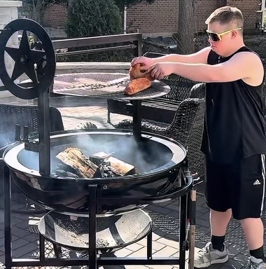 A boy wearing sunglasses and a black outfit places food on a raised grill over a fire pit outdoors. Smoke rises from the firewood below as he cooks in a backyard setting on a sunny day.
