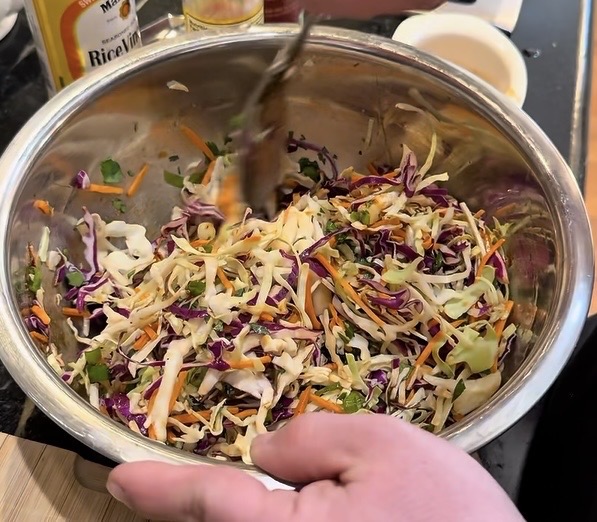 A hand mixing a colorful coleslaw salad with shredded cabbage, carrots, and herbs in a large metal bowl. Bottles and kitchen items are visible in the background.