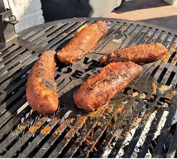 Four seasoned sausages are being grilled on a round barbecue grill, with char marks visible and some spice residue scattered on the grate. Sunlight casts shadows across the scene.
