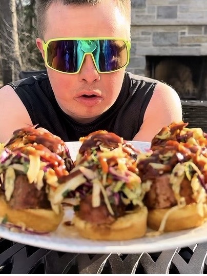 A person wearing large, reflective sunglasses holds a plate of loaded meatball sliders topped with slaw and sauce, with an outdoor fireplace in the background.