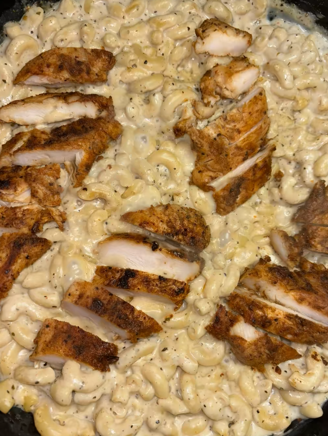 Close-up of creamy macaroni and cheese topped with sliced, seasoned grilled chicken breast pieces. The pasta is coated in a rich cheese sauce and the chicken appears juicy with a browned, spiced crust.