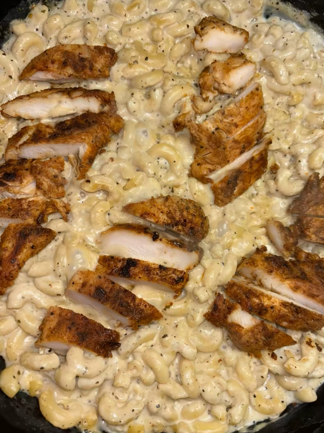 Close-up of creamy macaroni and cheese topped with sliced, seasoned grilled chicken breast in a pan. The pasta is coated in a rich cheese sauce, and the chicken is arranged on top.