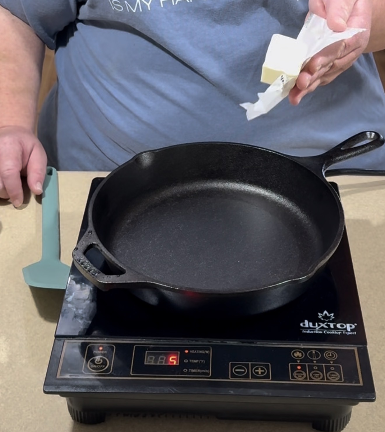 A person holding a stick of butter above a black cast iron skillet on an induction cooktop, preparing to cook. A blue spatula rests nearby on the counter.
