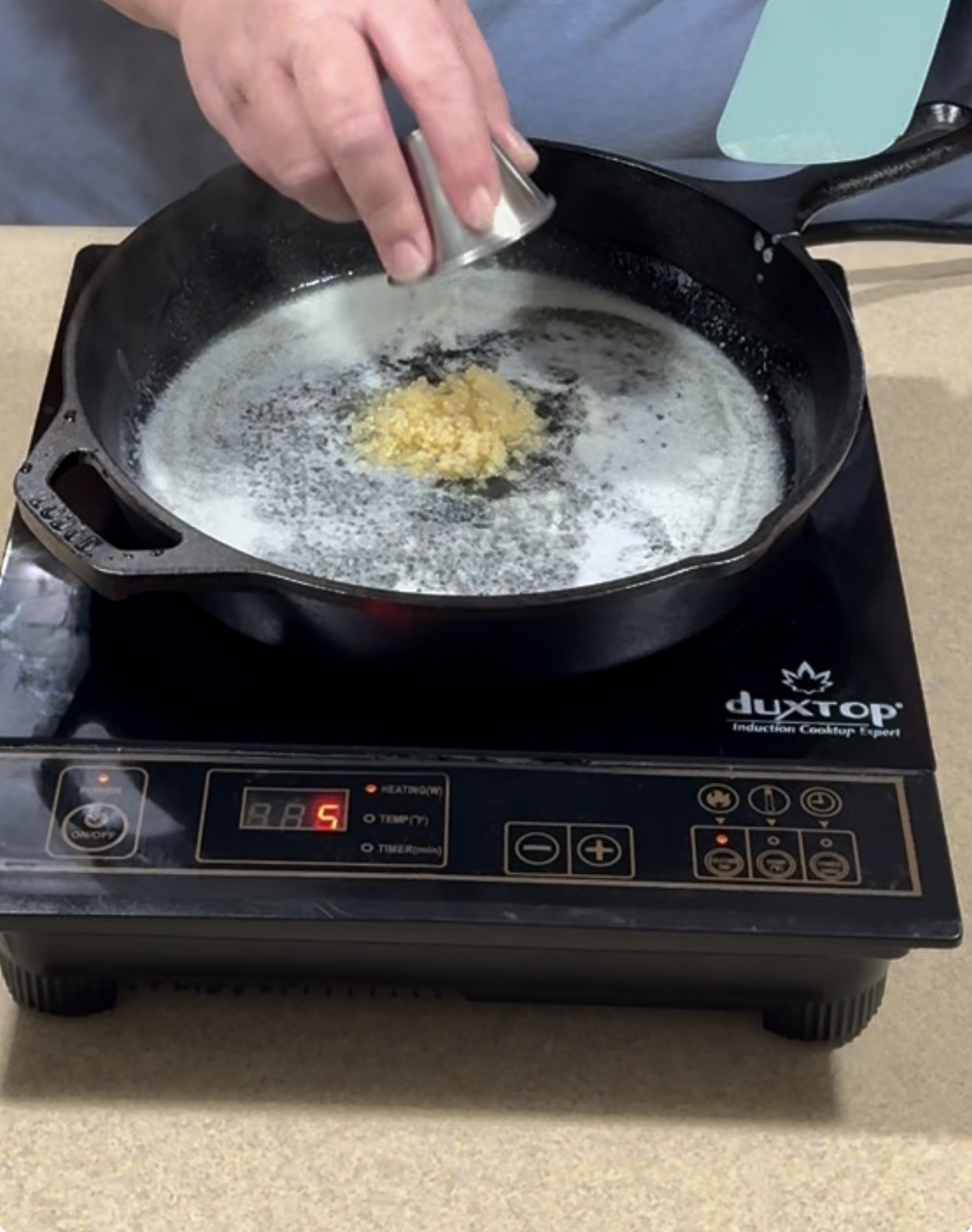 A person adds minced garlic from a small container into a cast iron skillet with melted butter on an induction cooktop.