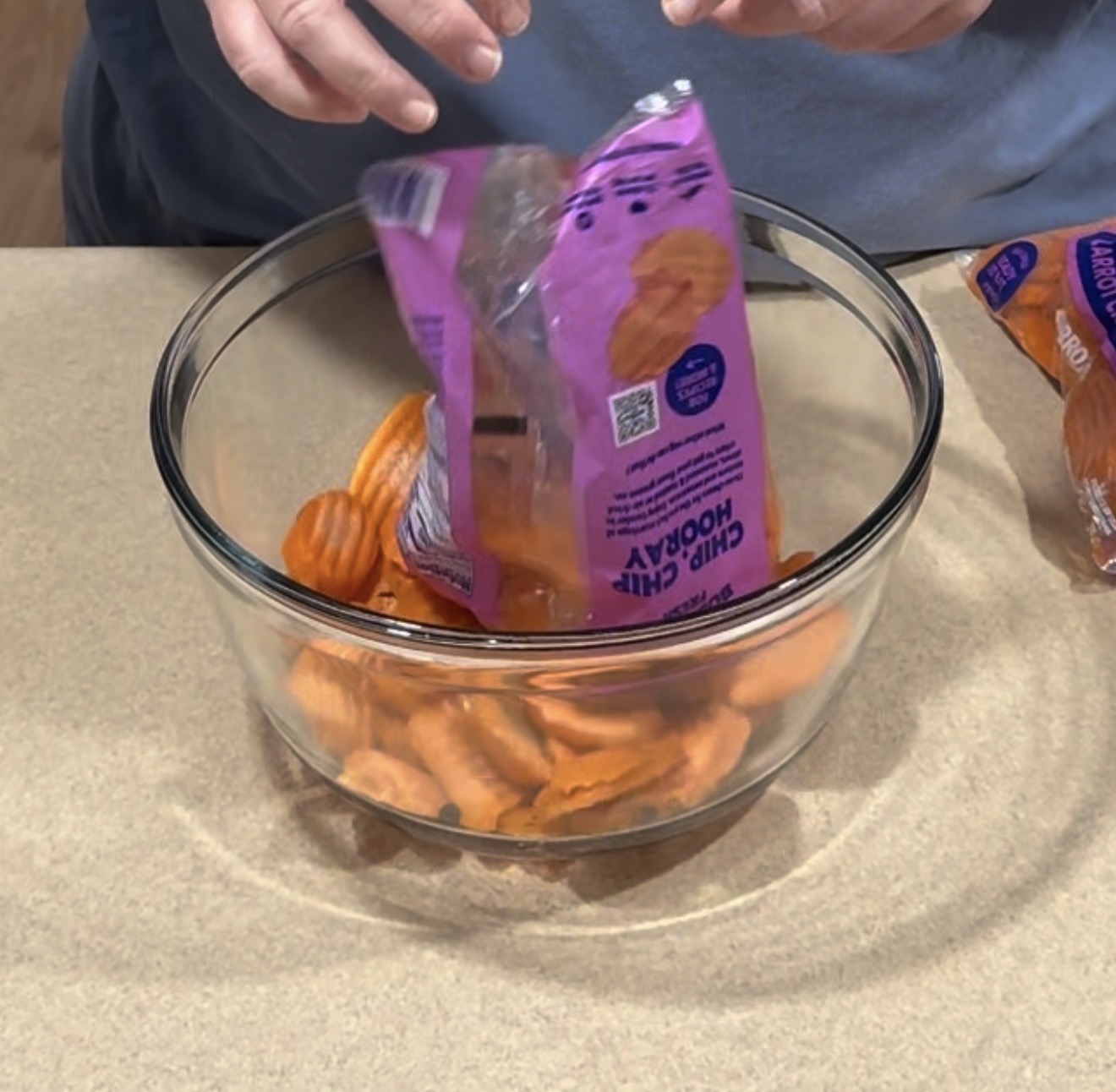 A person is pouring orange crinkle-cut chips from a purple bag into a clear glass bowl on a beige countertop.