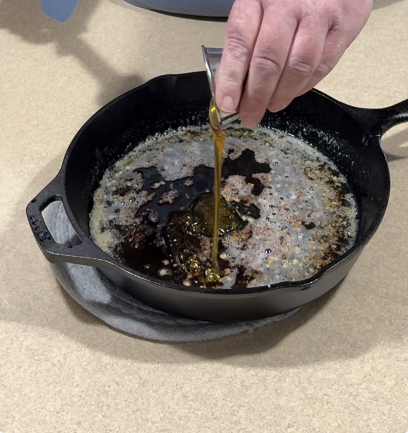 A hand pours a small metal cup of honey or syrup into a black cast iron skillet with bubbling melted butter on a countertop.
