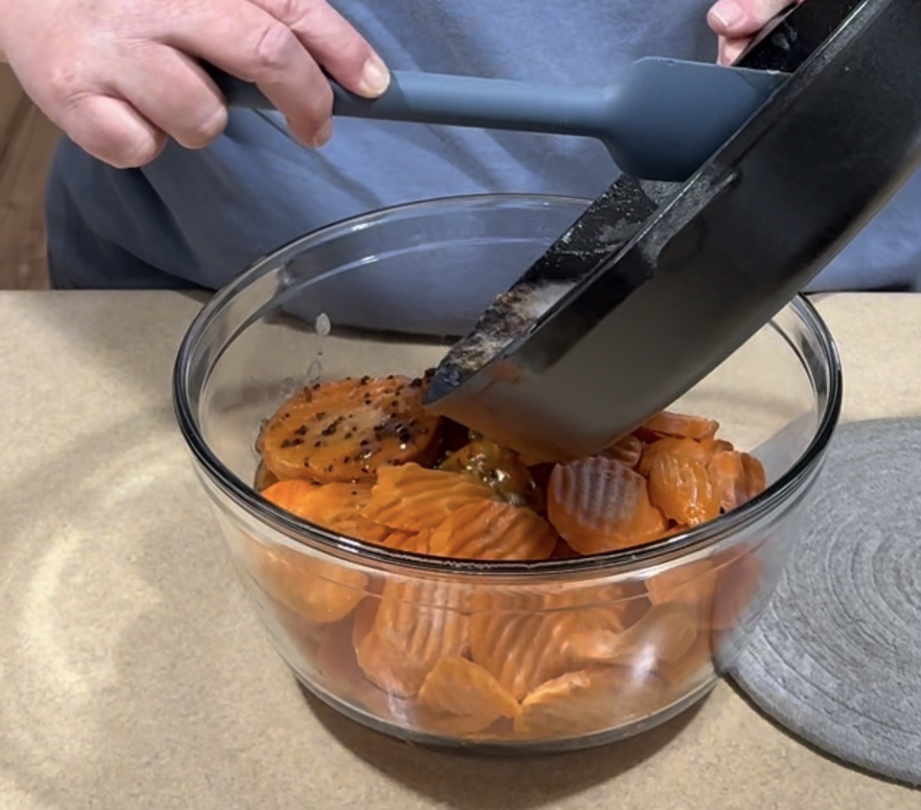 A person pours a mixture from a black pan into a glass bowl filled with sliced cooked carrots using a blue spatula. The bowl is on a beige countertop.