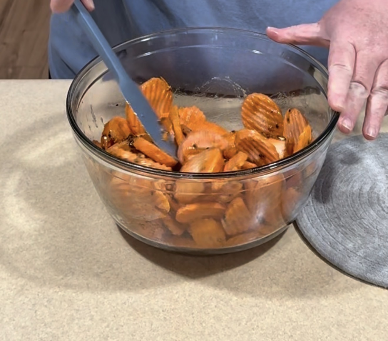 A person mixes crinkle-cut sweet potato slices in a glass bowl with a blue spatula on a beige countertop.