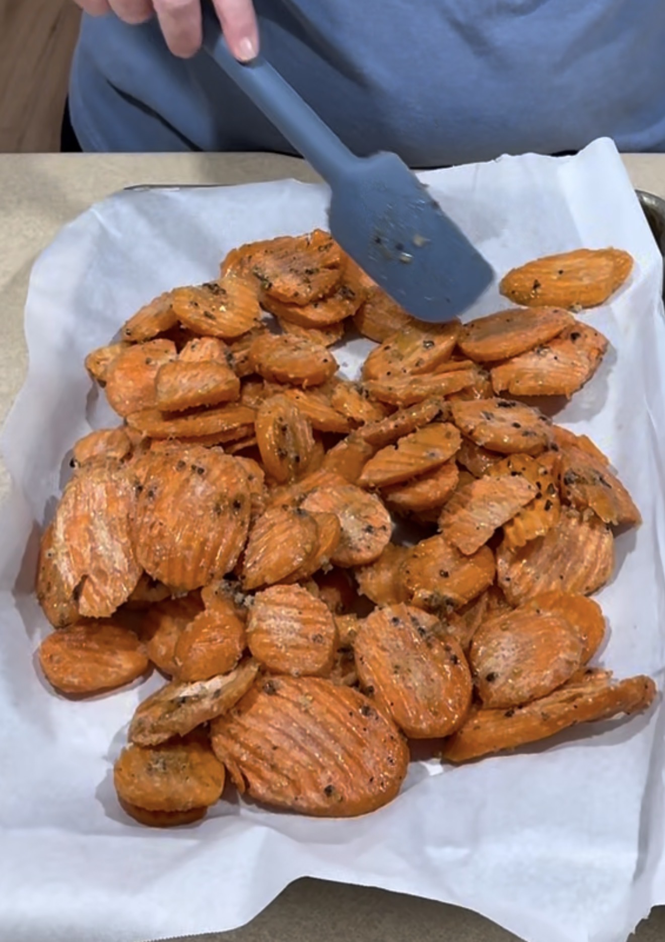 A person uses a blue spatula to spread out seasoned, crinkle-cut roasted carrots on a baking sheet lined with parchment paper.