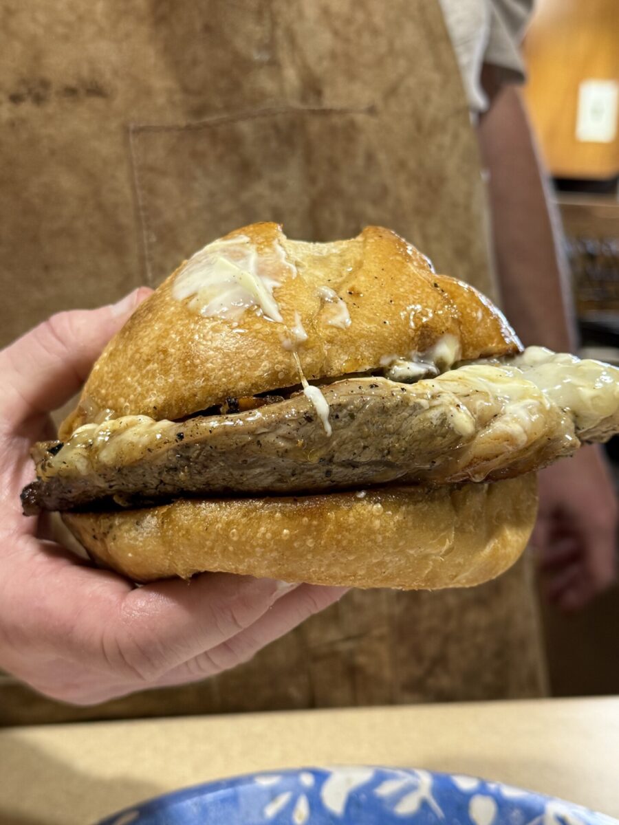 A hand holds a sandwich with a thick slice of meat and melted cheese inside a golden, toasted bun. The background shows a person wearing a brown apron.