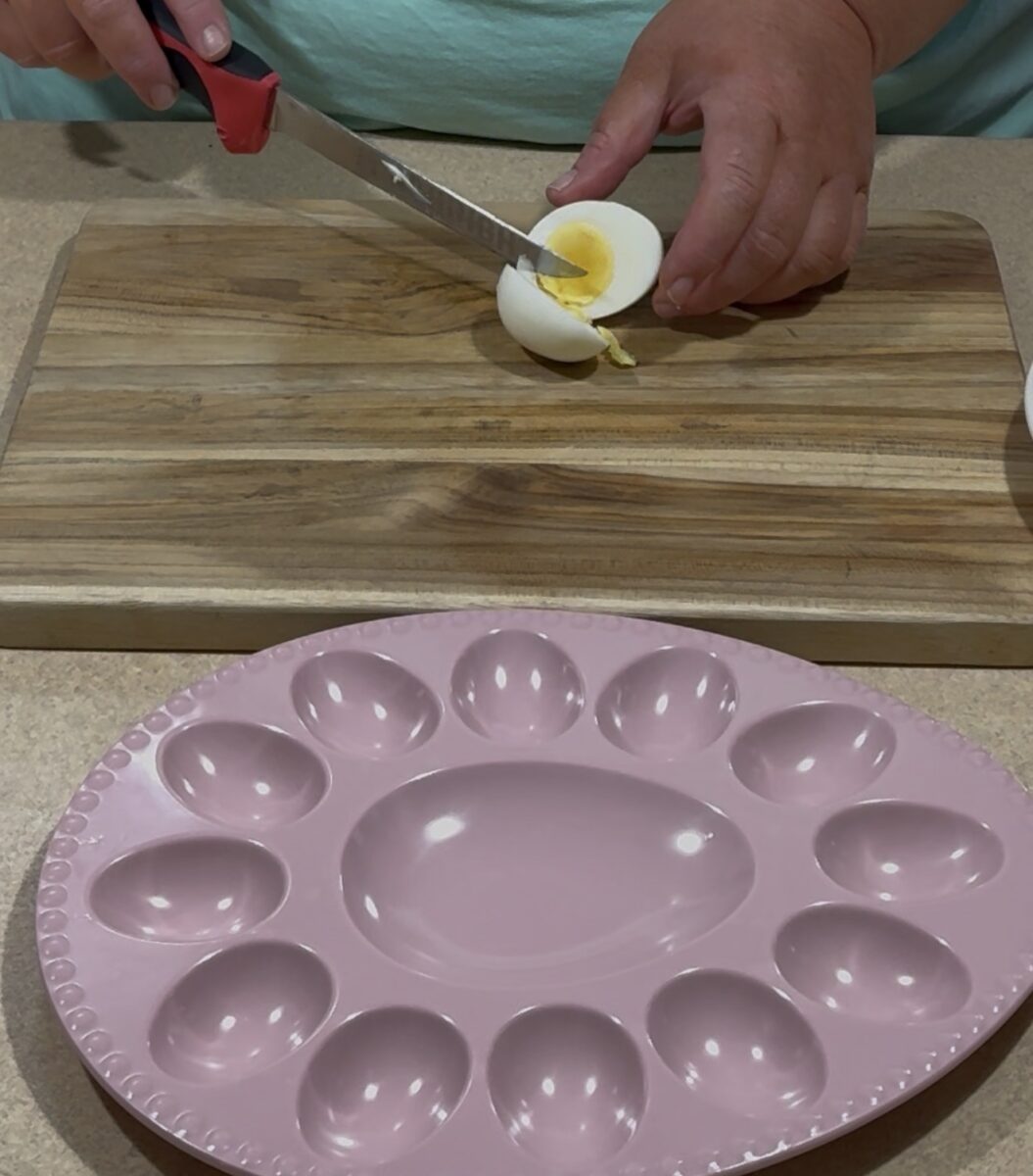 A person is slicing a hard-boiled egg on a wooden cutting board. In front of them is a pink egg tray with spaces for deviled eggs, sitting on a beige countertop.