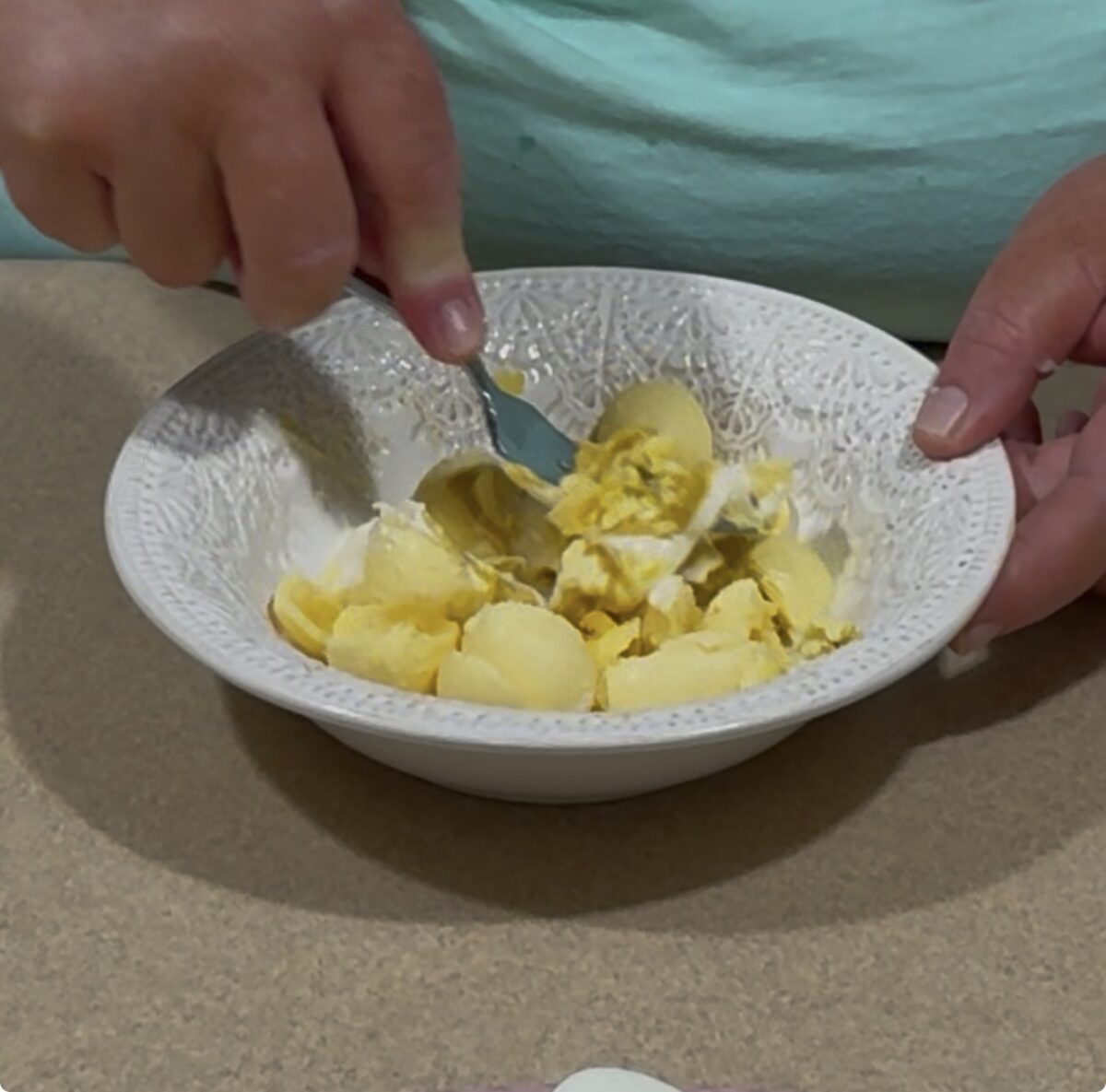 A person is mashing hard-boiled eggs with a fork in a white textured bowl on a countertop.