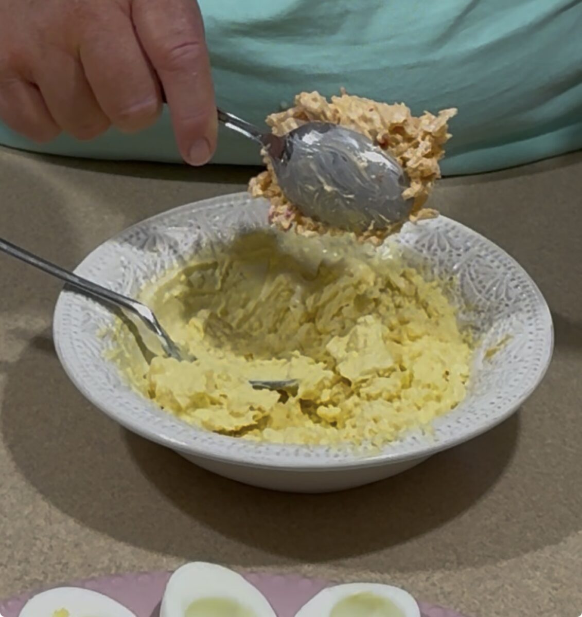 A person holds a spoonful of yellow deviled egg filling over a bowl with more filling. Halved hard-boiled eggs are visible on a plate in the foreground.