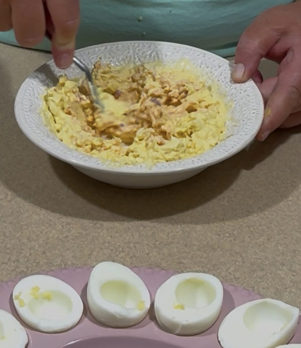 A person mixes a creamy yellow filling in a bowl with a fork. In the foreground, halved hard-boiled eggs with the yolks removed are arranged on a plate, ready to be filled.