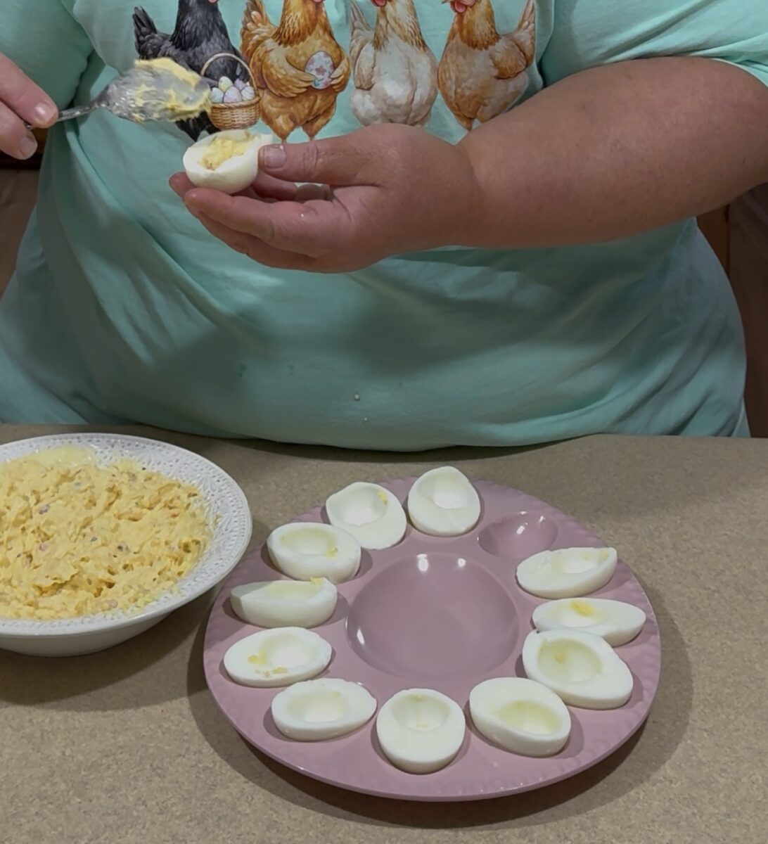 A person wearing a shirt with a chicken design is making deviled eggs, spooning filling into egg white halves arranged on a pink tray, with a bowl of egg yolk mixture nearby.