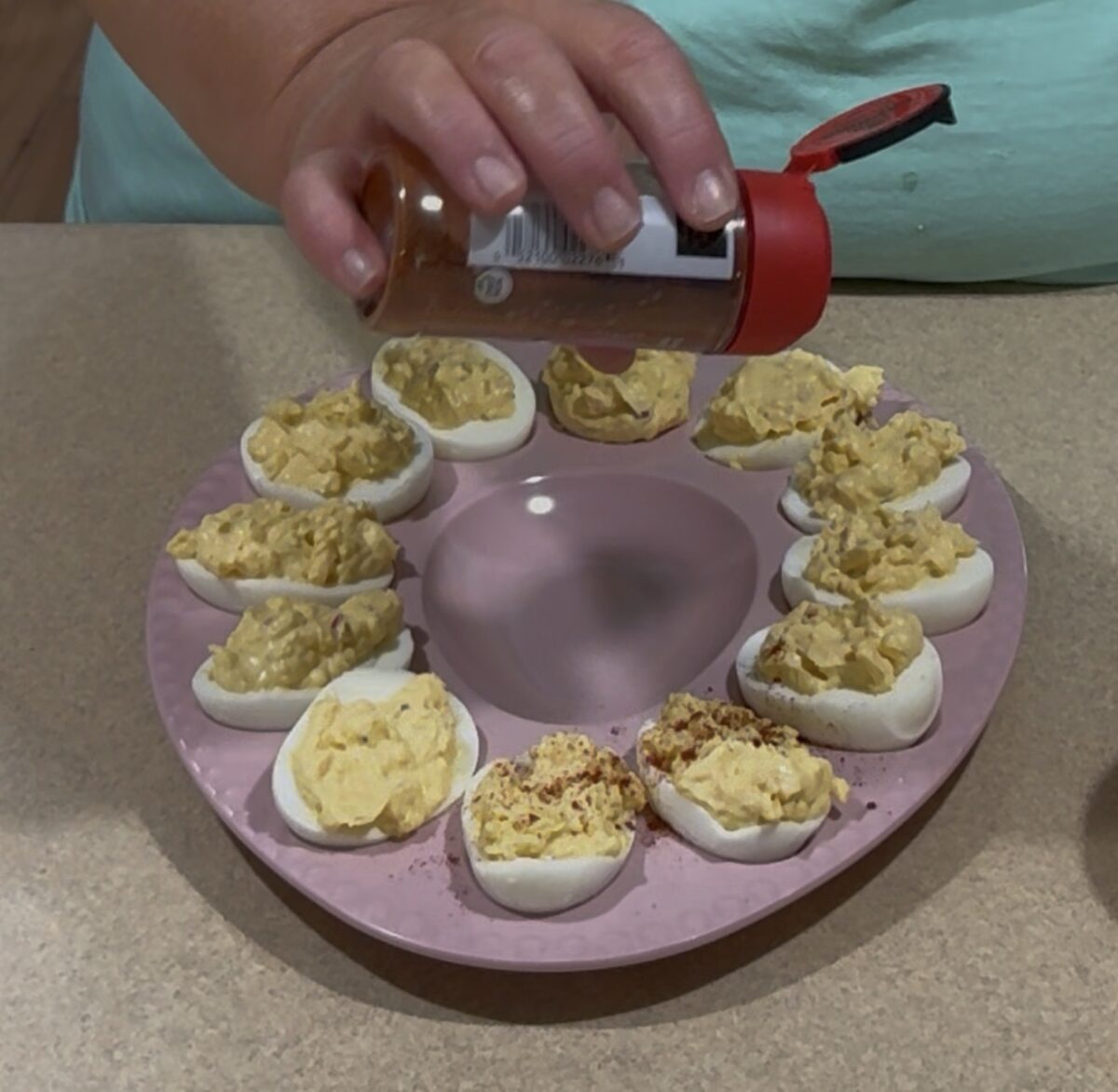 A person sprinkles seasoning onto deviled eggs arranged in a circle on a pink plate, with their hand holding a spice container above the eggs.
