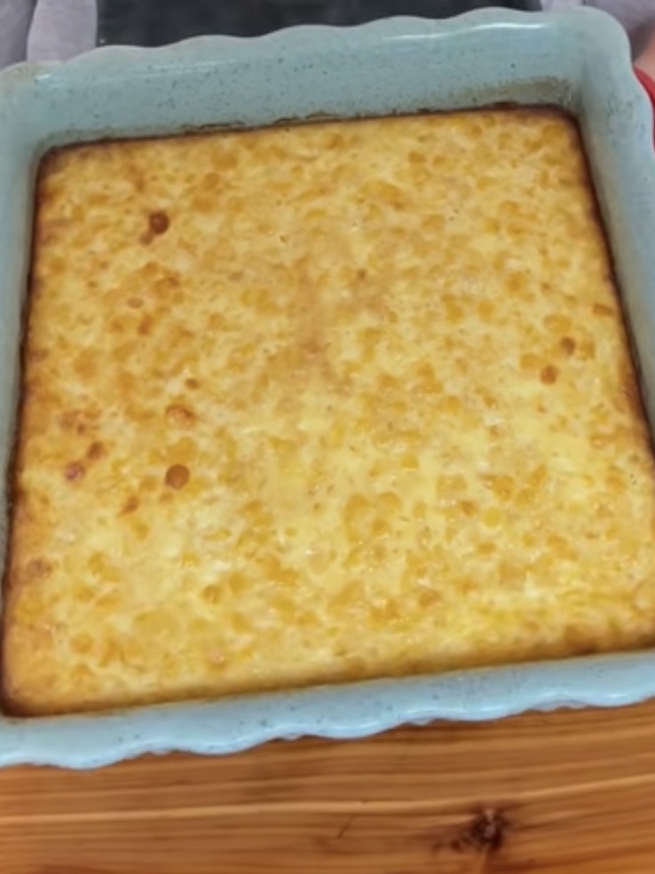 A close-up of a golden-brown baked corn pudding in a blue square baking dish, sitting on a wooden surface. The dessert has a slightly uneven, bubbly top.