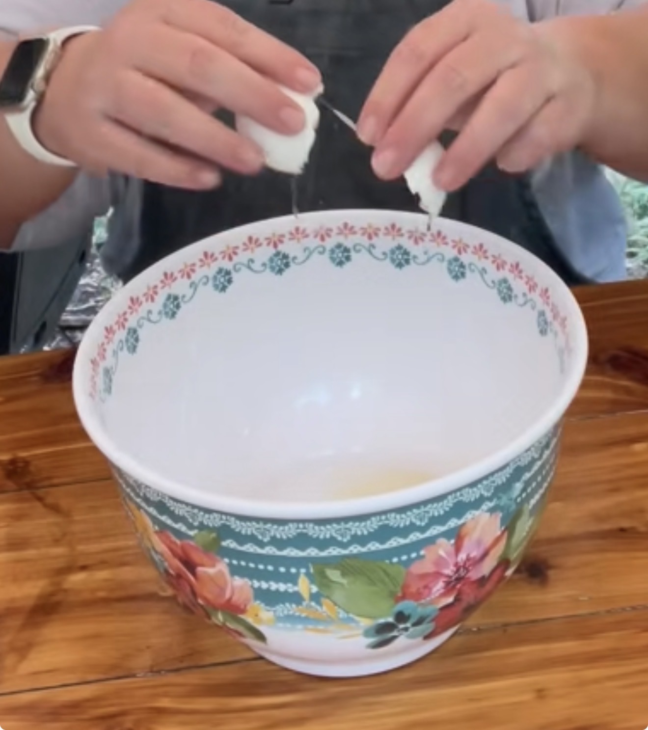 A person cracks an egg into a large, floral-patterned mixing bowl on a wooden table.