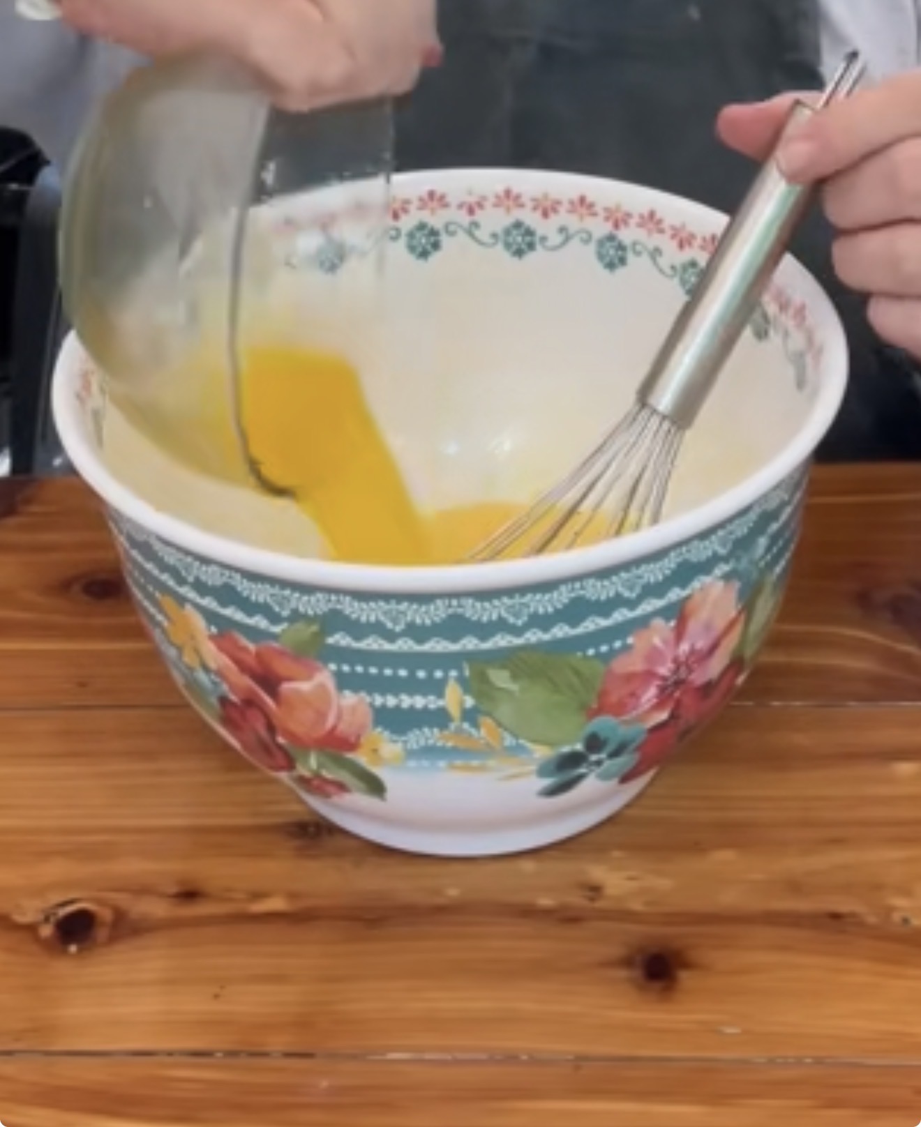 A person pours a bowl of beaten eggs into a large floral-patterned mixing bowl on a wooden surface, holding a whisk in one hand.