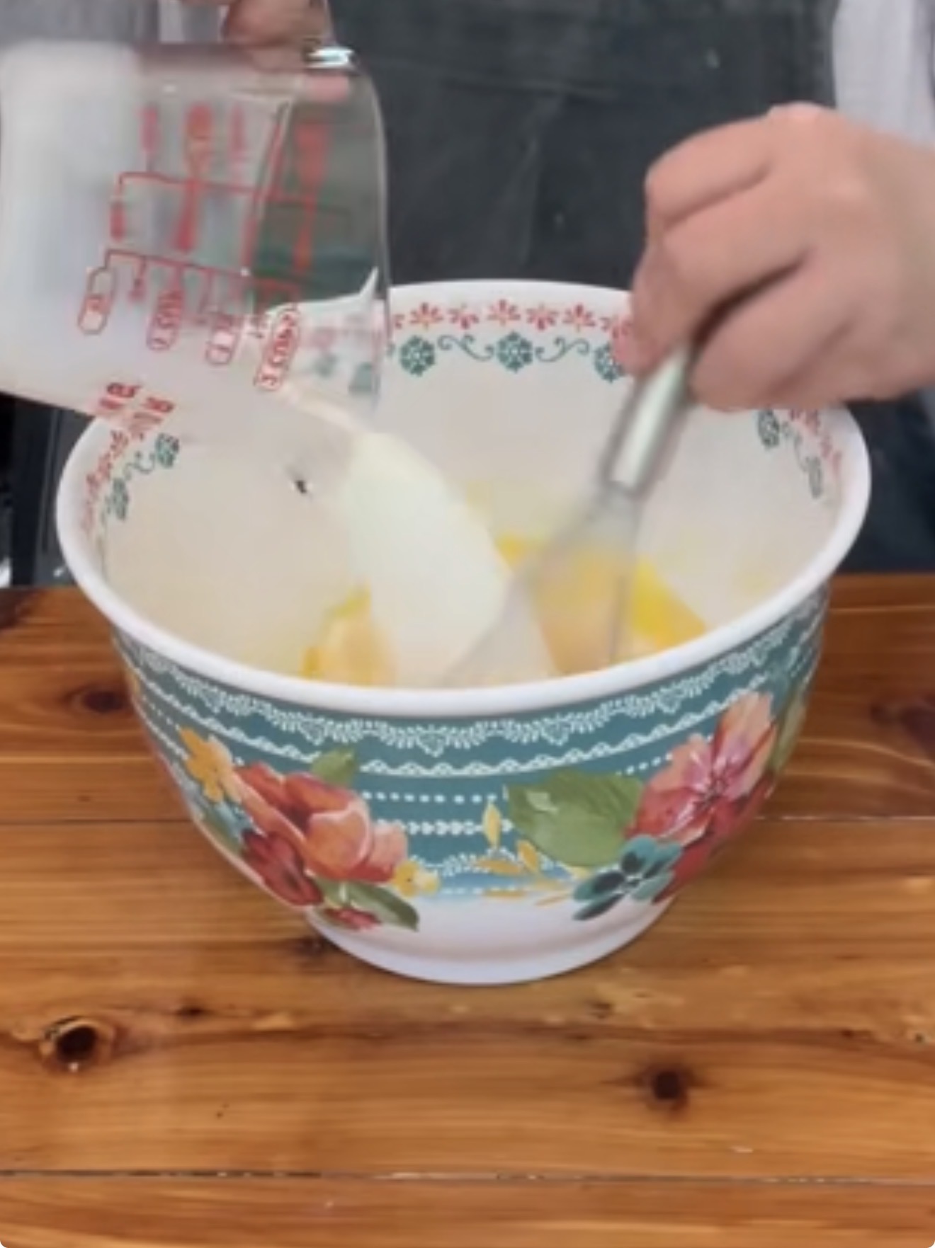 A person pours milk from a measuring cup into a patterned mixing bowl while whisking eggs, preparing ingredients on a wooden table.
