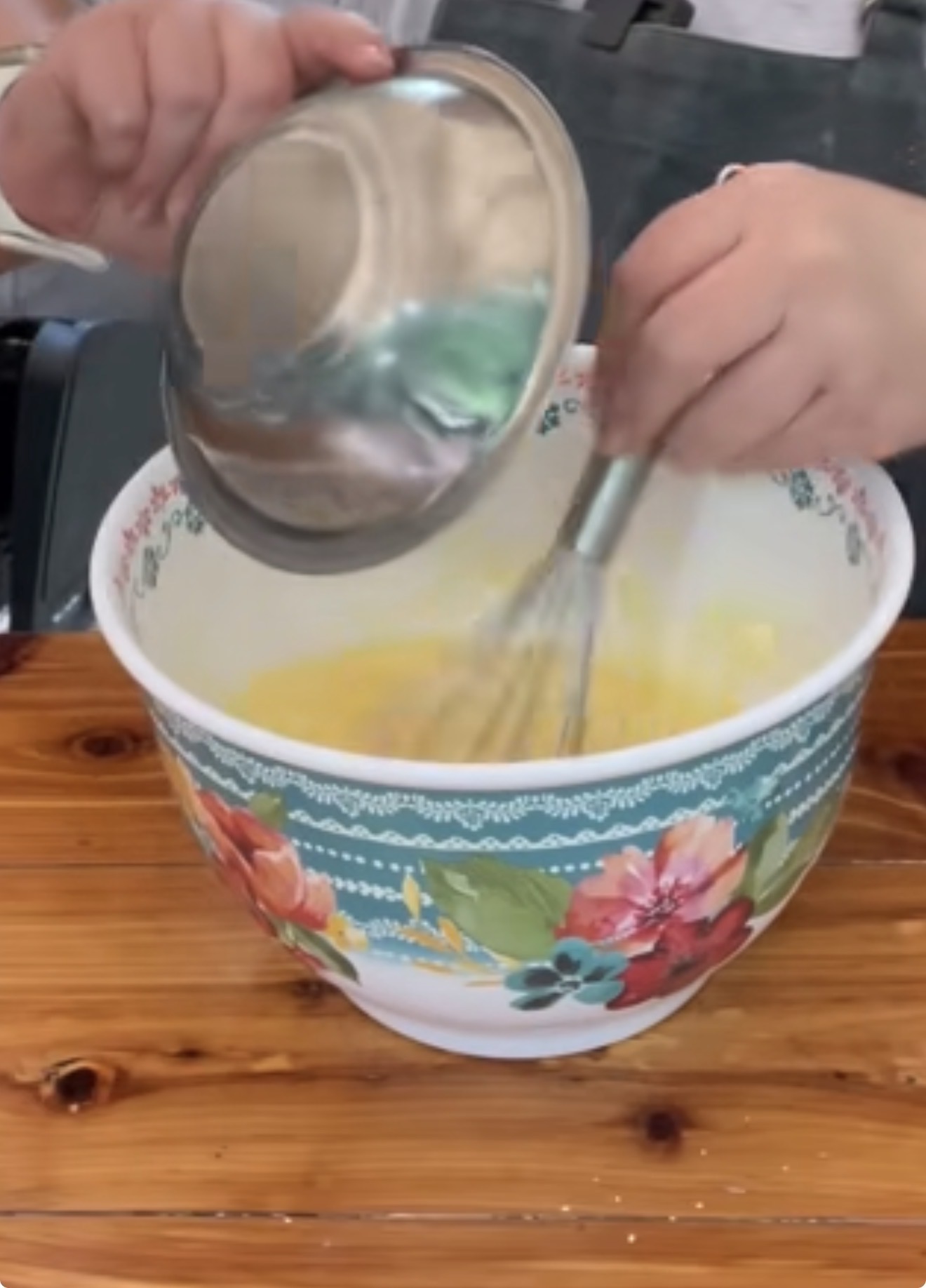 A person is whisking ingredients in a large, floral-patterned mixing bowl on a wooden table while holding a metal bowl lid in one hand.