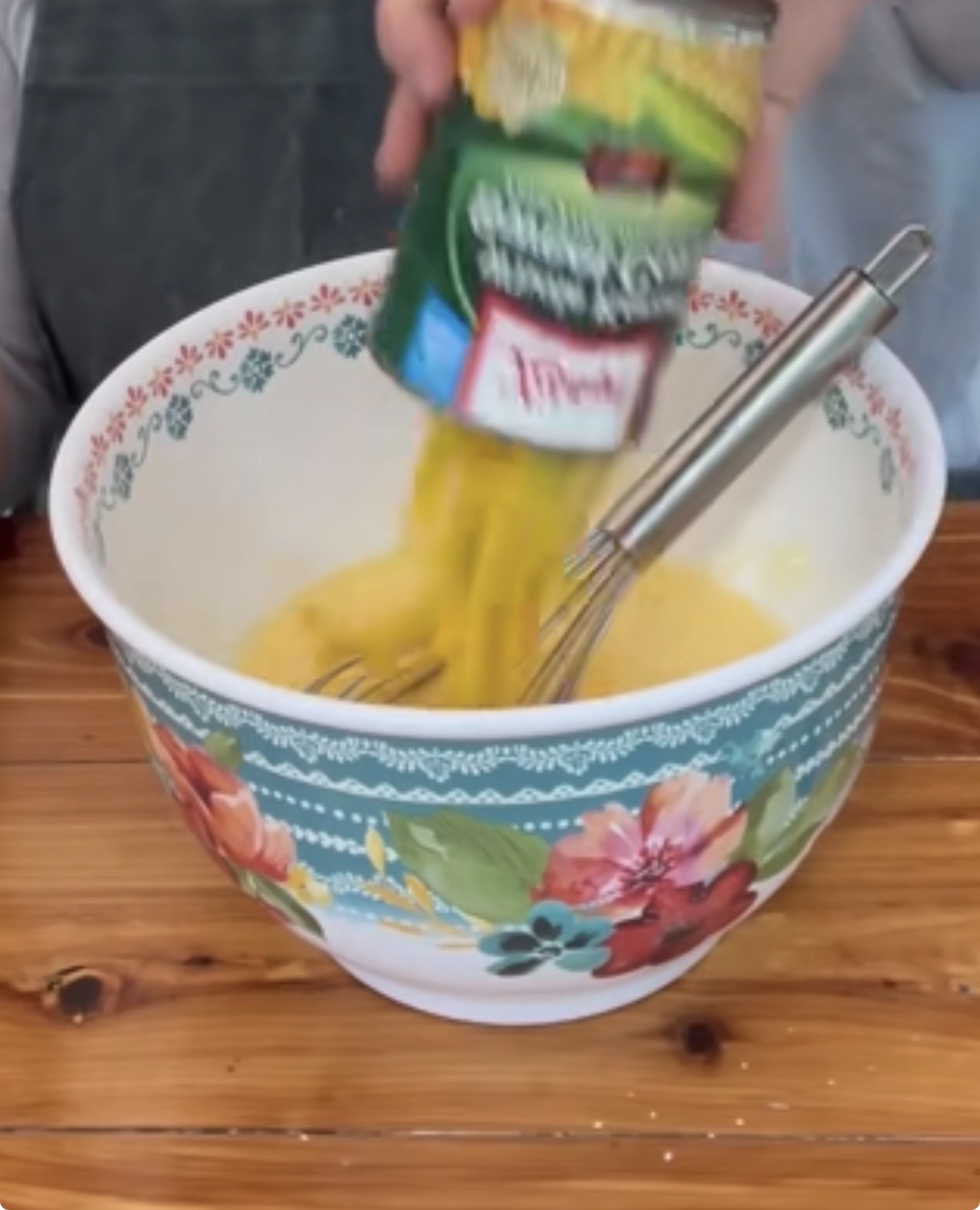 A can of cut green beans is being poured into a mixing bowl with a whisk and a yellow mixture. The floral-patterned bowl sits on a wooden table.