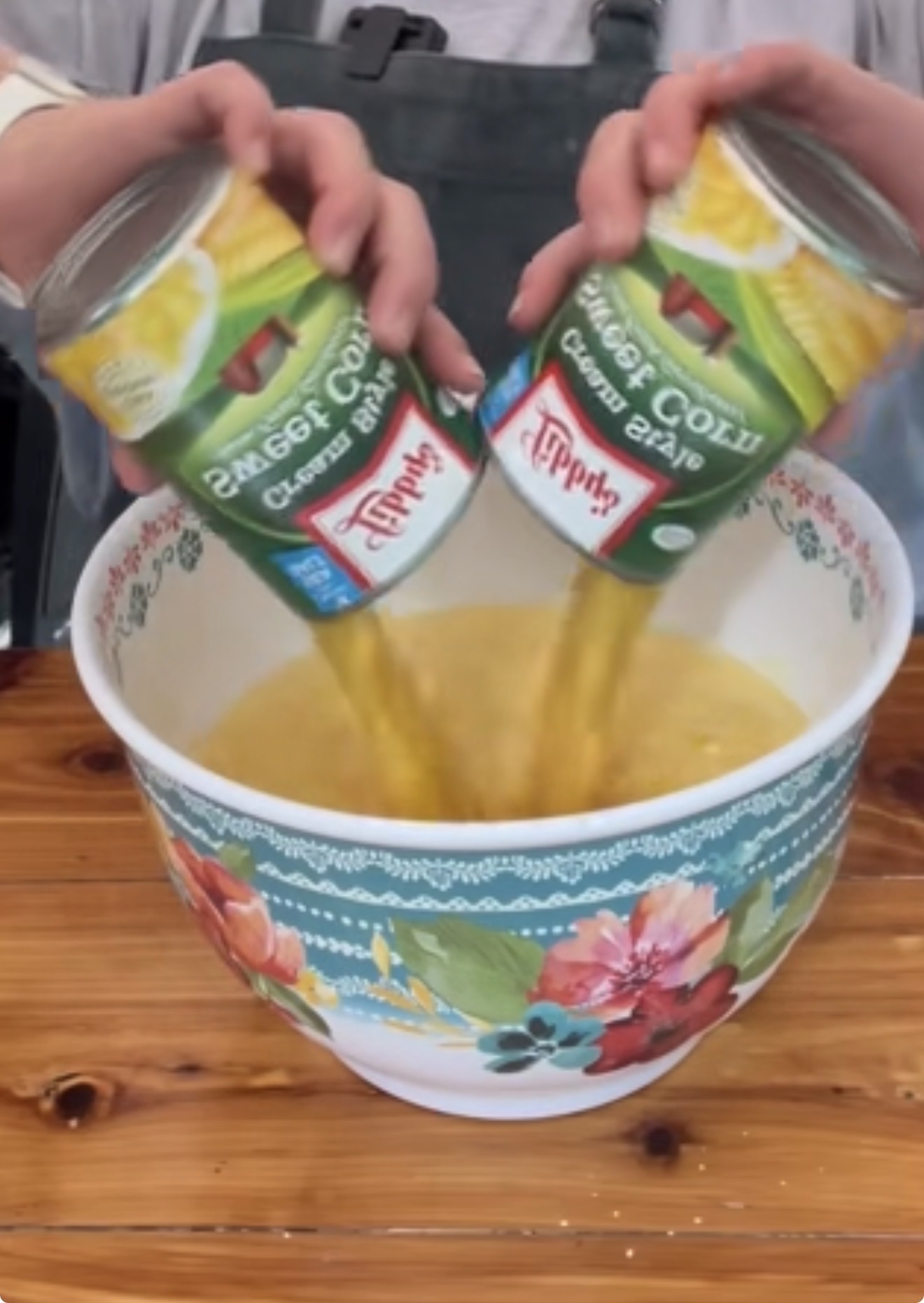 A person pours two cans of Tippins Cream Style Golden Sweet Corn into a large floral-patterned mixing bowl on a wooden table.