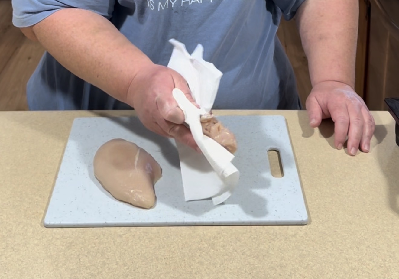 A person using a paper towel to pat dry a raw chicken breast on a white cutting board, with another chicken breast beside it on a kitchen counter.