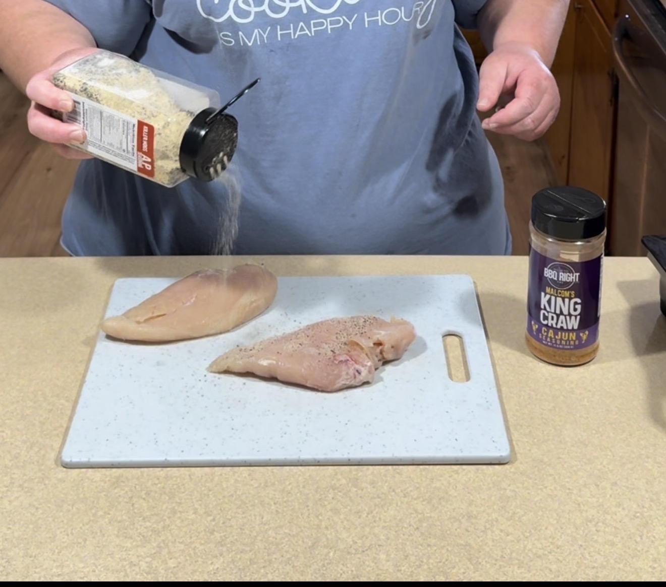 A person sprinkles seasoning from a container onto raw chicken breasts on a white cutting board, with another seasoning jar labeled “King Craw Cajun” placed nearby on the counter.