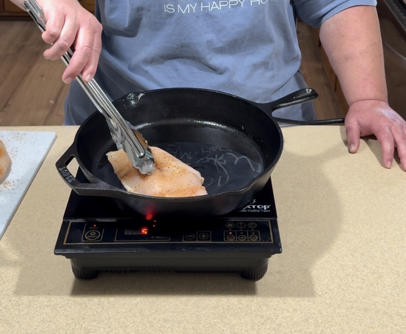 A person uses tongs to place a seasoned fish fillet into a black cast iron skillet on an electric stovetop. Another hand rests on the counter, and a plate with more fish is nearby.