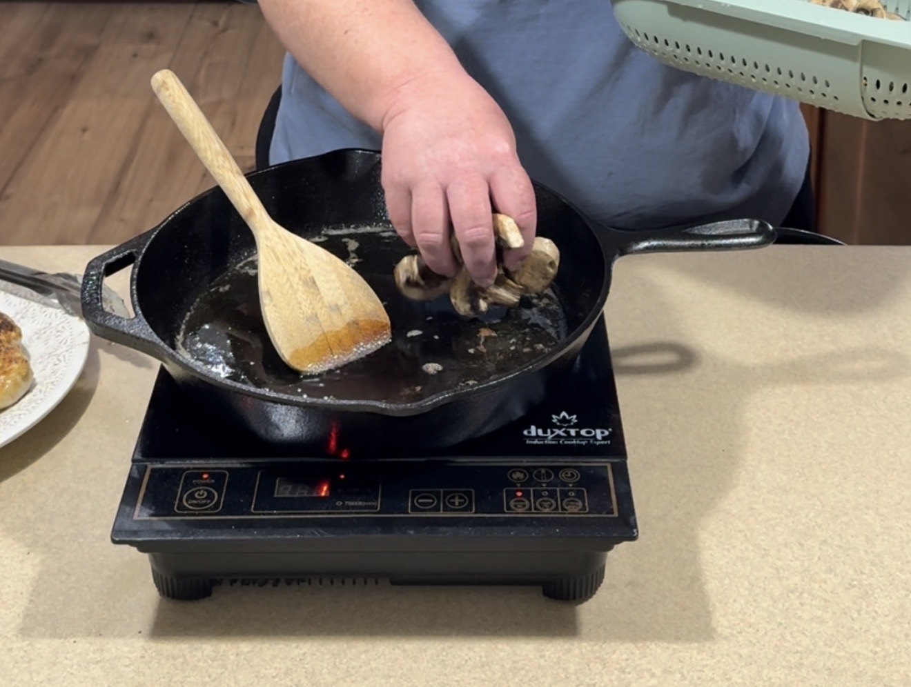 A person adds sliced mushrooms to a black cast iron skillet on an induction cooktop, using a wooden spoon to stir. A light green colander with more mushrooms is visible nearby.