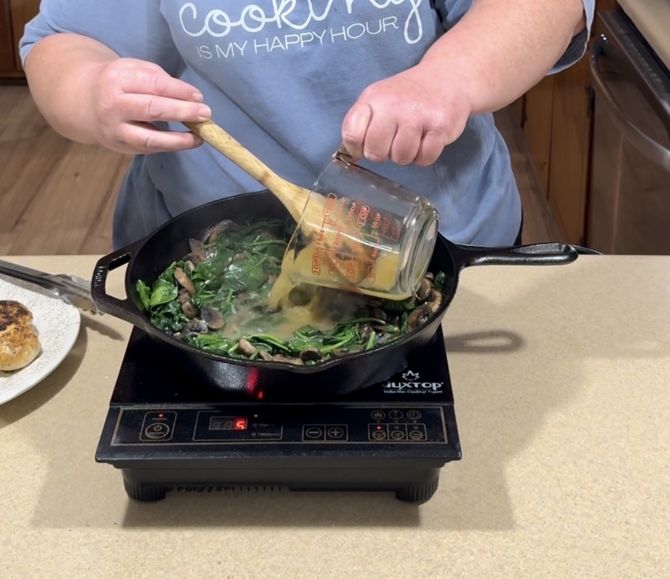 A person in a blue shirt pours broth from a measuring cup into a cast iron skillet with mushrooms and spinach, cooking on an induction cooktop in a kitchen.