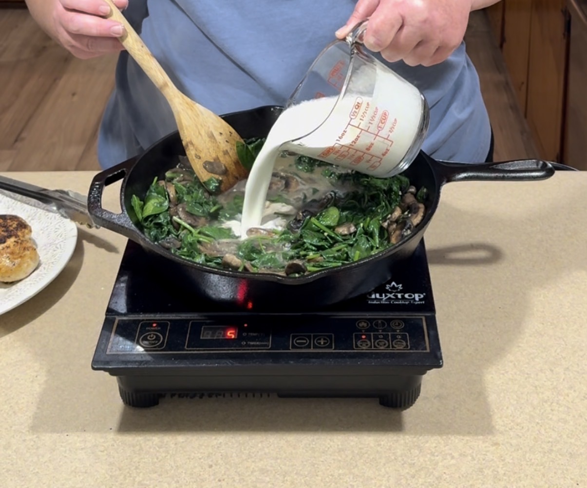 A person pours milk from a measuring cup into a skillet with sautéed greens and mushrooms while stirring with a wooden spoon on an electric cooktop.