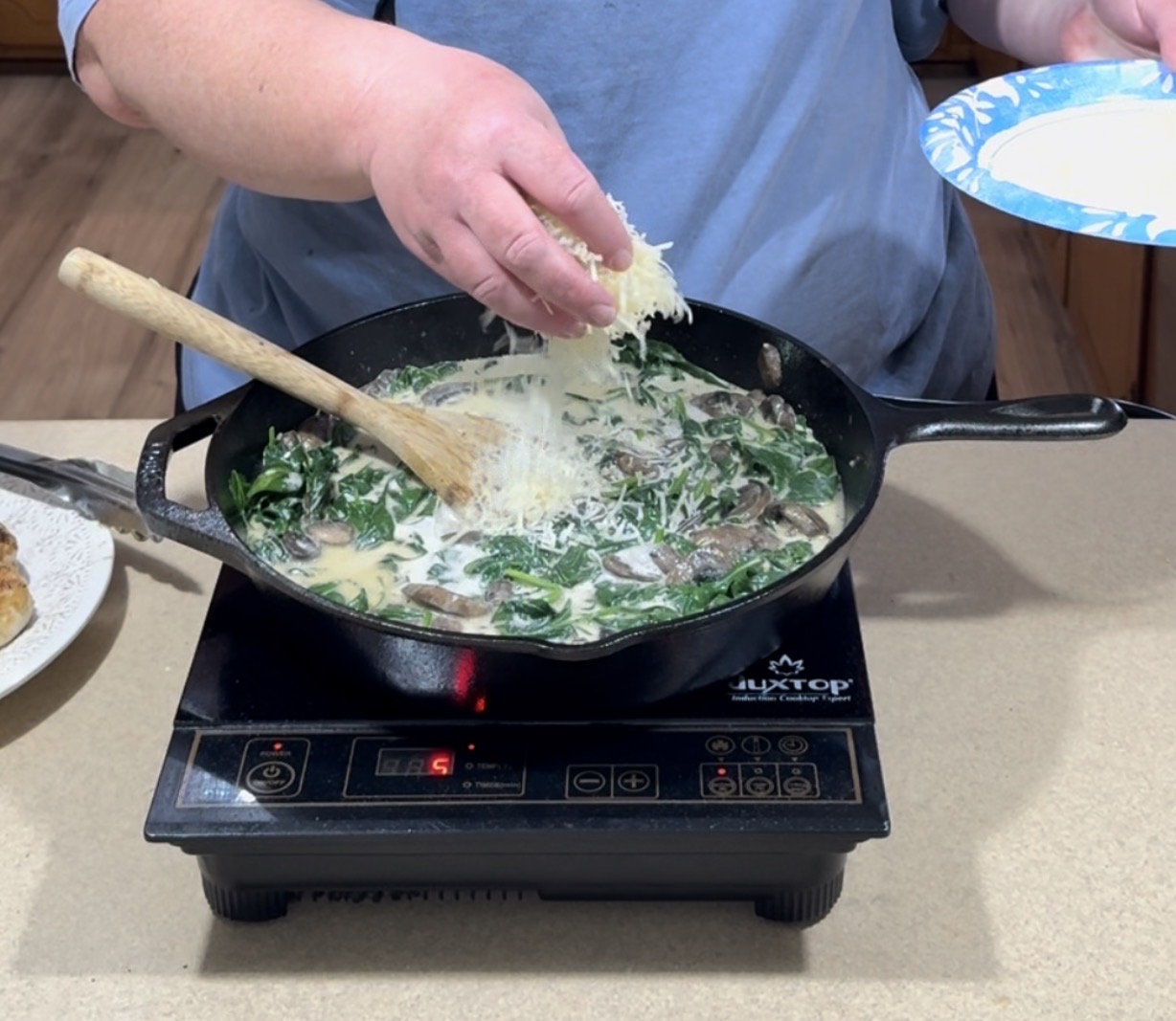 A person sprinkles shredded cheese into a skillet filled with spinach, mushrooms, and a creamy sauce on an electric cooktop, using a wooden spoon to stir.