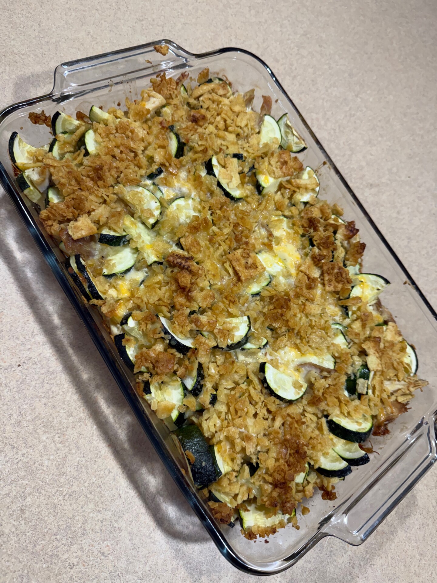 A glass baking dish filled with a baked casserole featuring sliced zucchini and yellow squash, topped with a golden, crispy cracker or breadcrumb crust, sitting on a beige countertop.