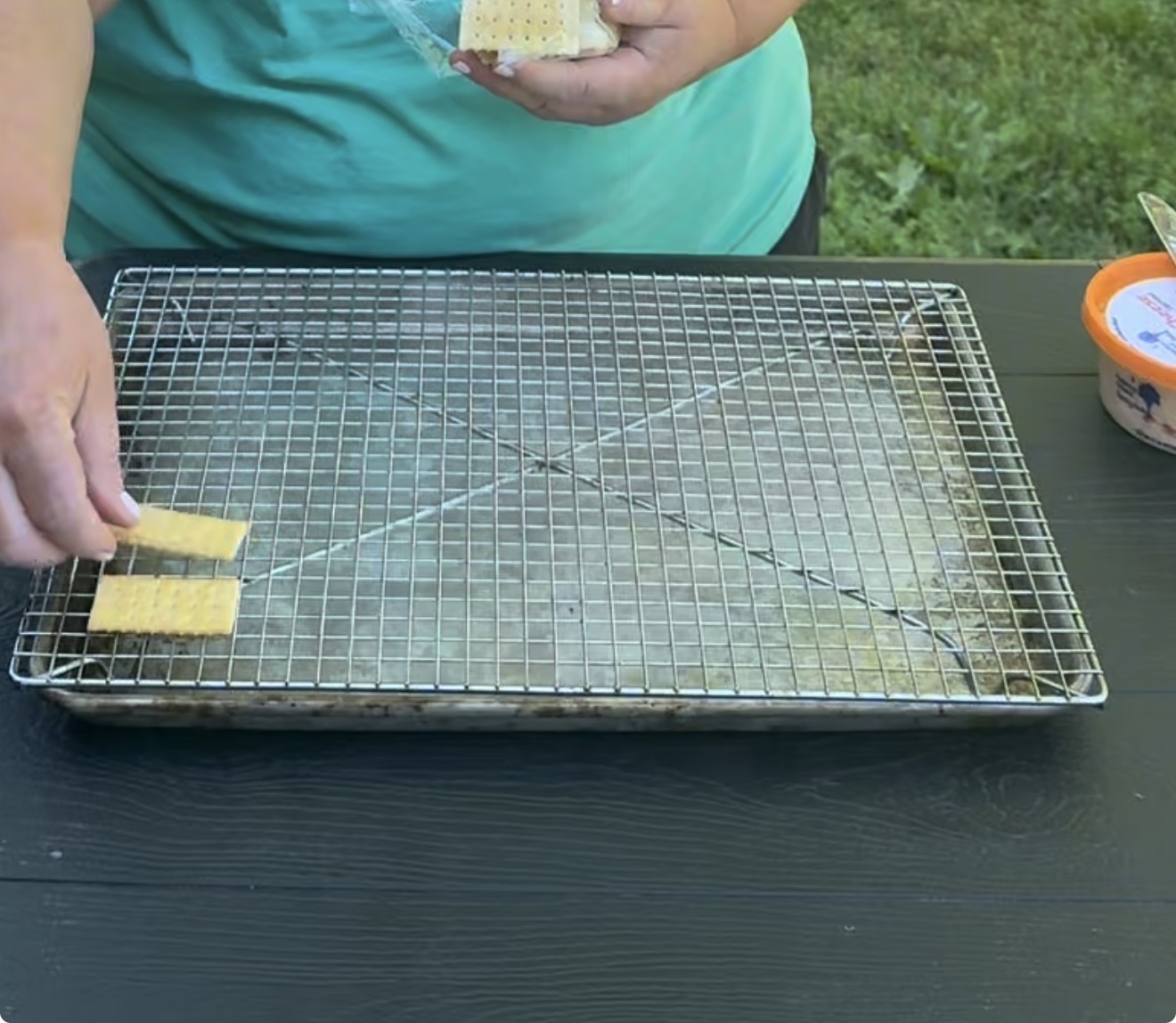 A person places a rectangular cracker on a wire rack set over a baking sheet outdoors. Another cracker and a tub of dip or spread are visible on the table.