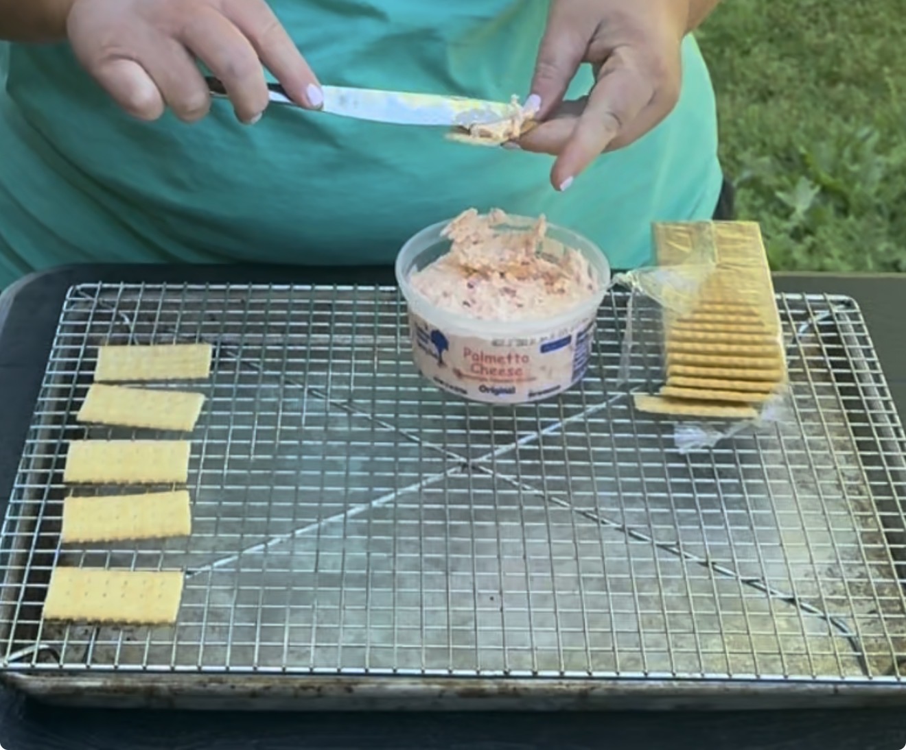 A person spreads pimento cheese onto a cracker over a cooling rack with several crackers and a container of pimento cheese on it, outdoors on a grassy background.