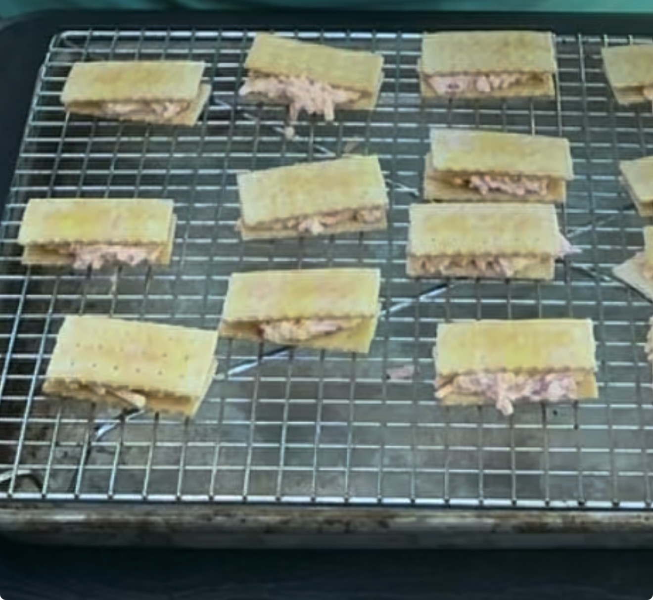Rectangular crackers with a pinkish filling, arranged in rows on a wire rack over a baking tray. The filling appears spread between two crackers, resembling small sandwiches.
