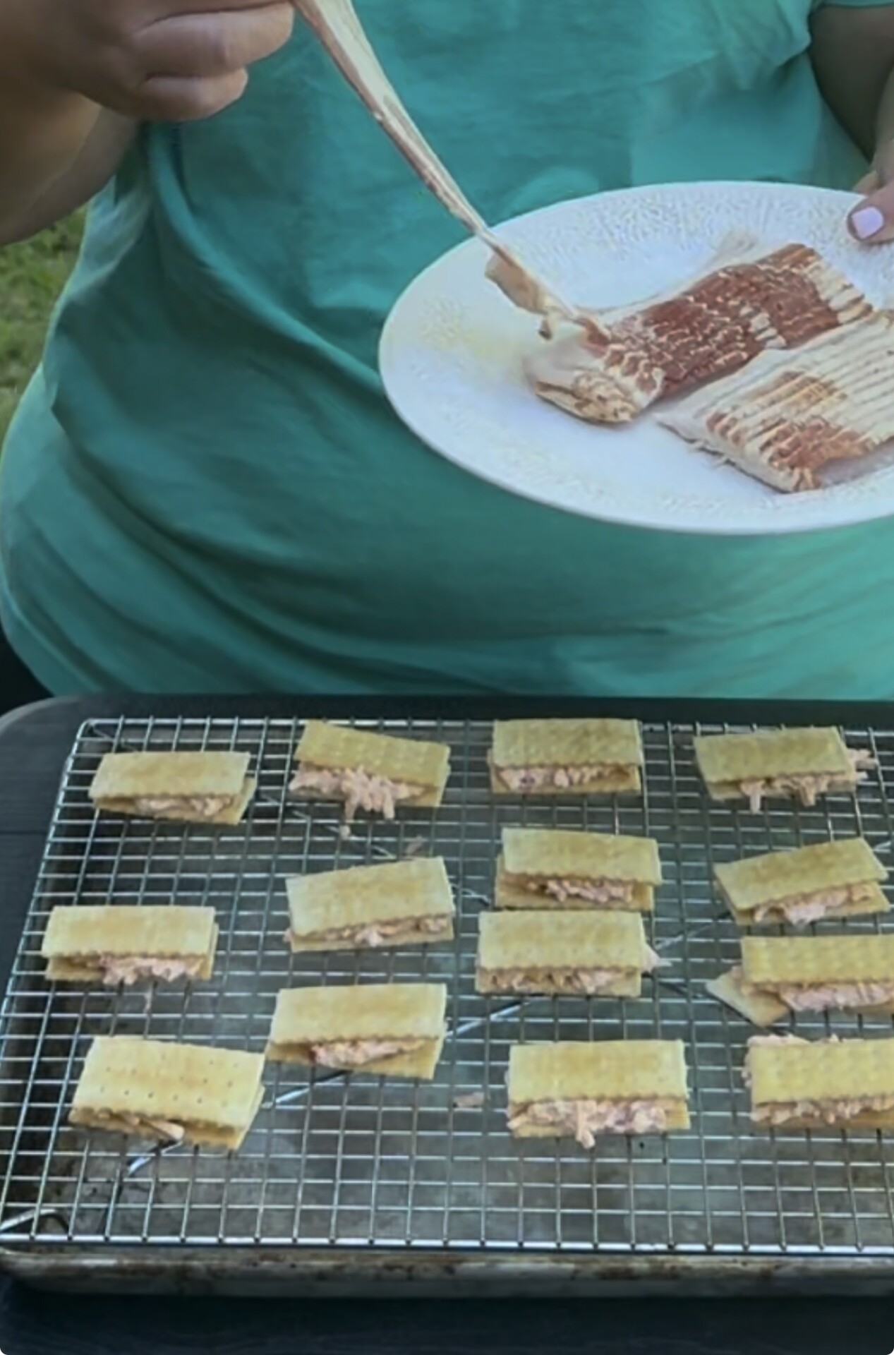 A person in a teal shirt wraps strips of bacon around small rectangular sandwiches on a wire rack, preparing them for cooking. A plate with more bacon strips is held in one hand.