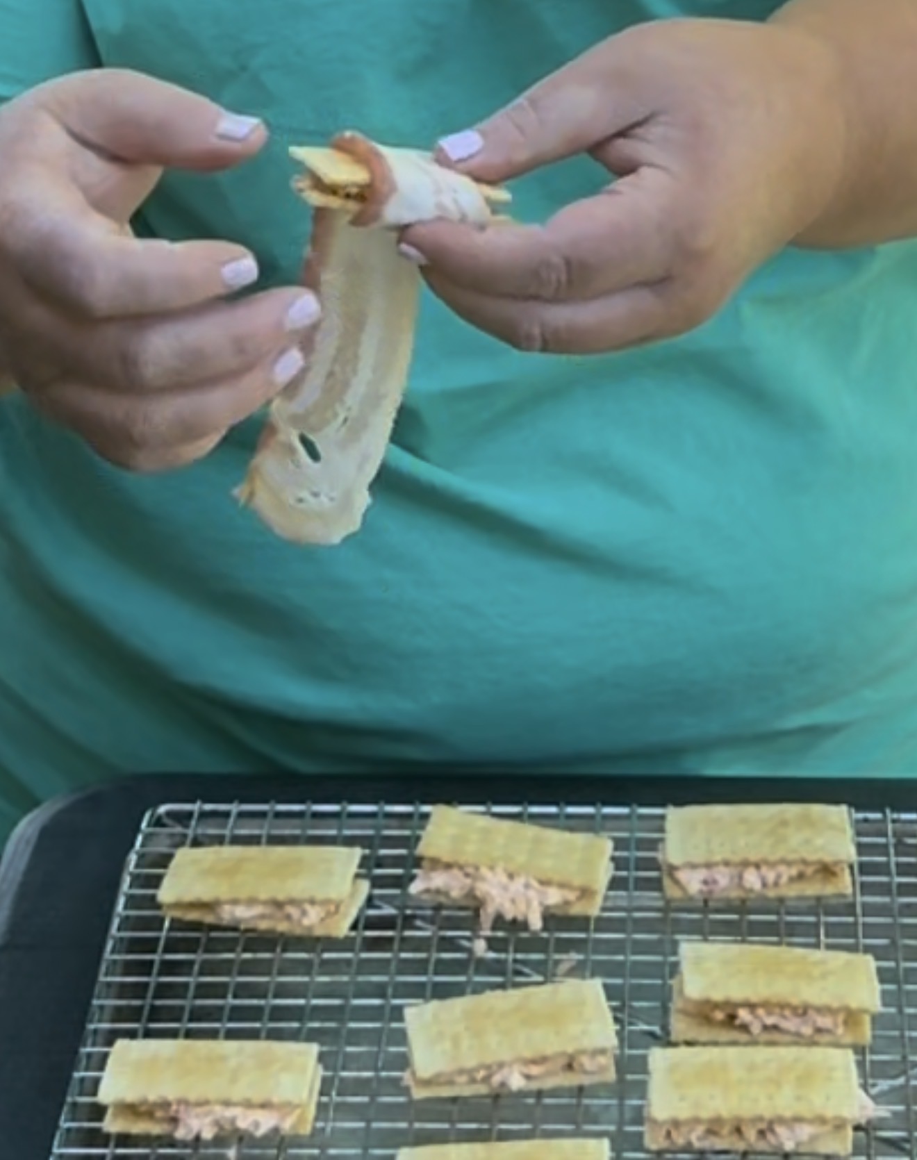 A person in a teal shirt holds a piece of bacon while assembling small sandwiches filled with a pinkish spread on a cooling rack.