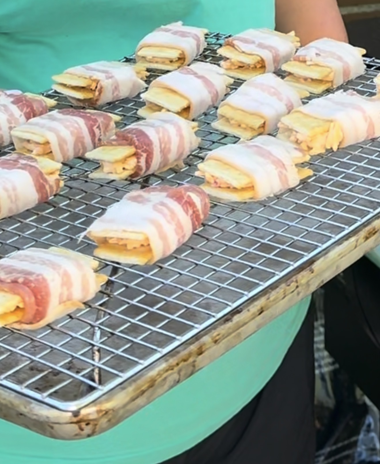 A person holding a metal rack filled with uncooked bacon-wrapped snacks, each containing layers of cheese and other ingredients, ready to be baked or grilled.