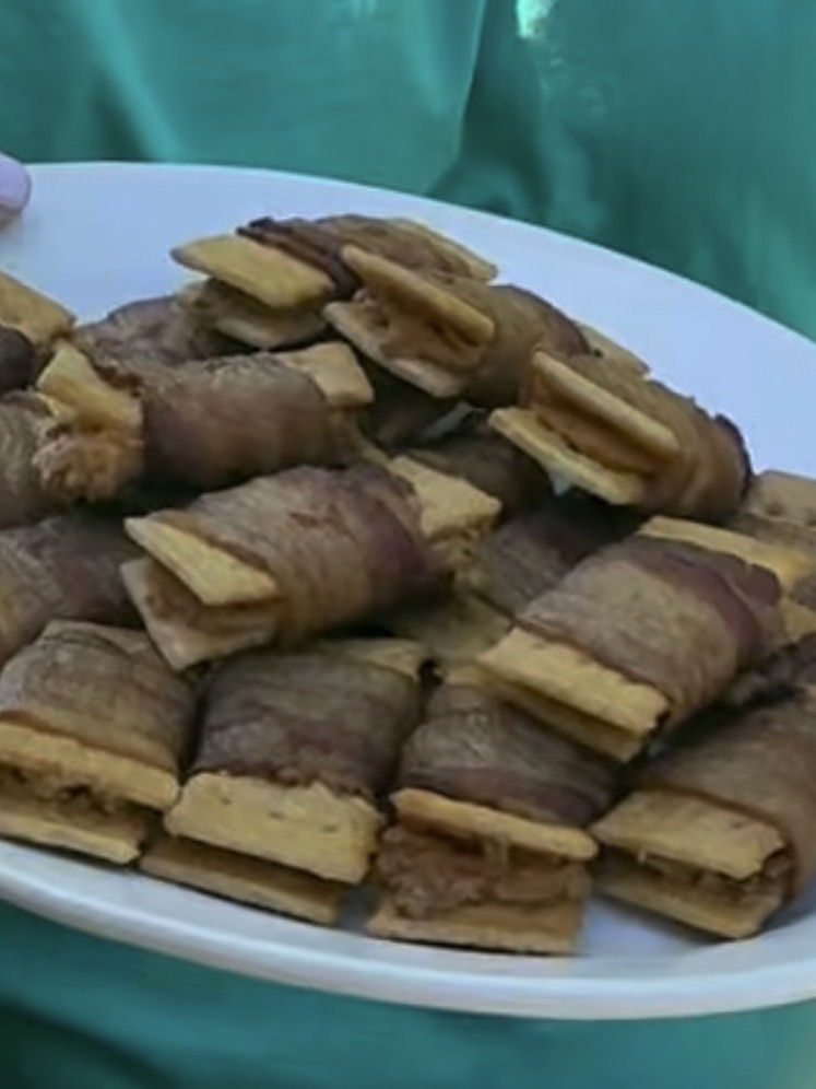 A white plate filled with rectangular, golden-brown snacks wrapped in bacon, held by someone wearing a green shirt.