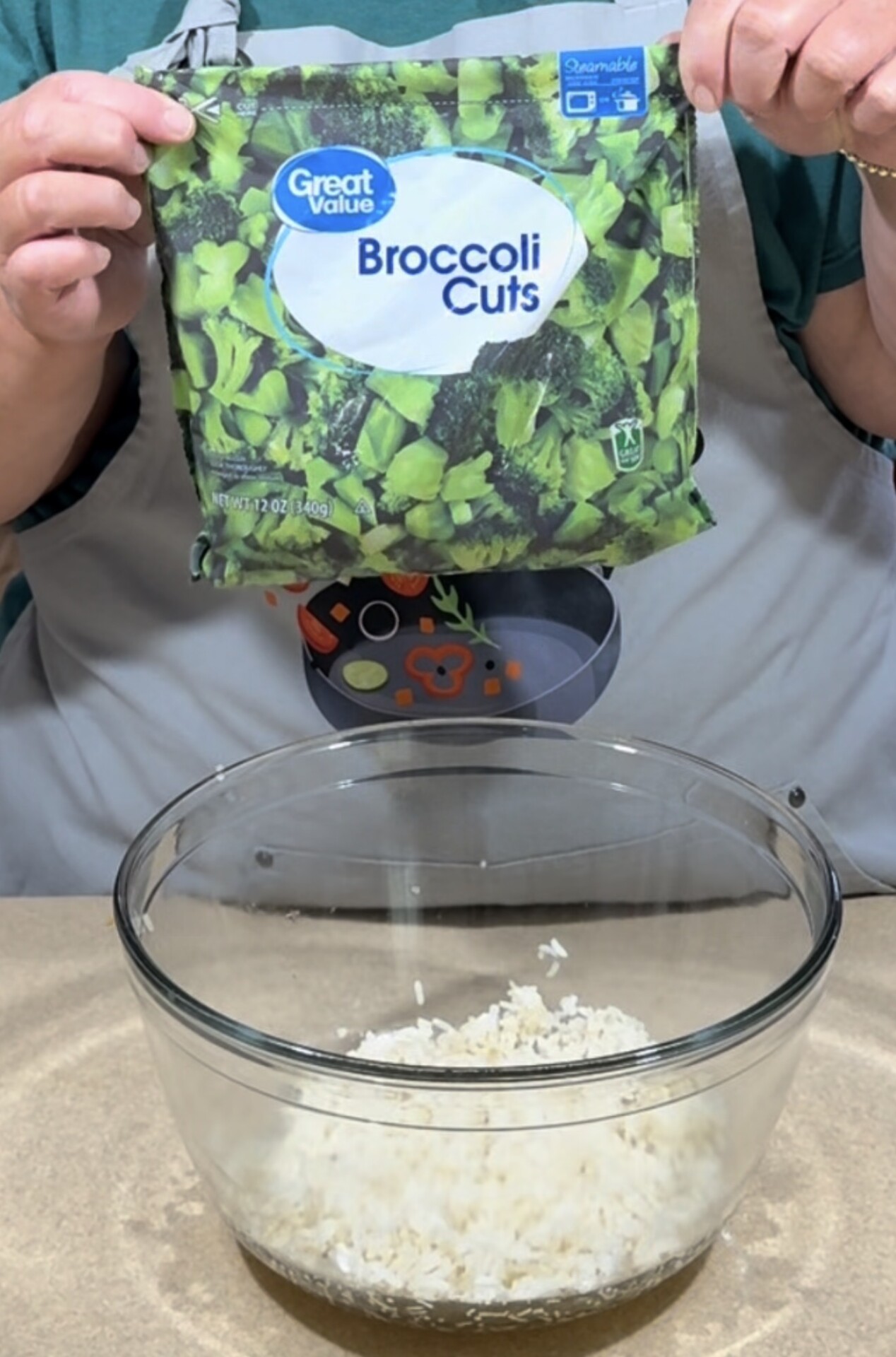 A person holds a bag of Great Value Broccoli Cuts above a clear glass bowl containing shredded white cheese or rice on a kitchen counter.