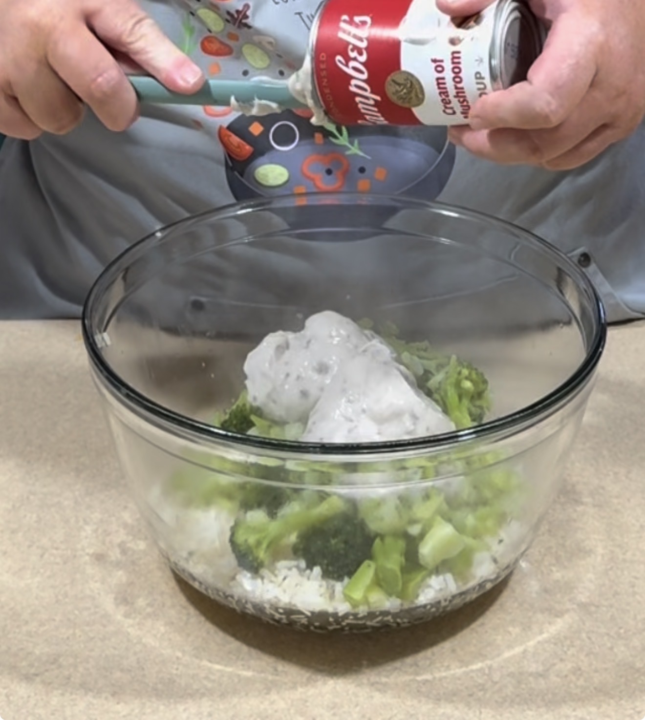 A person pours Campbells cream of mushroom soup from a can into a glass bowl containing broccoli, rice, and other ingredients on a beige countertop.