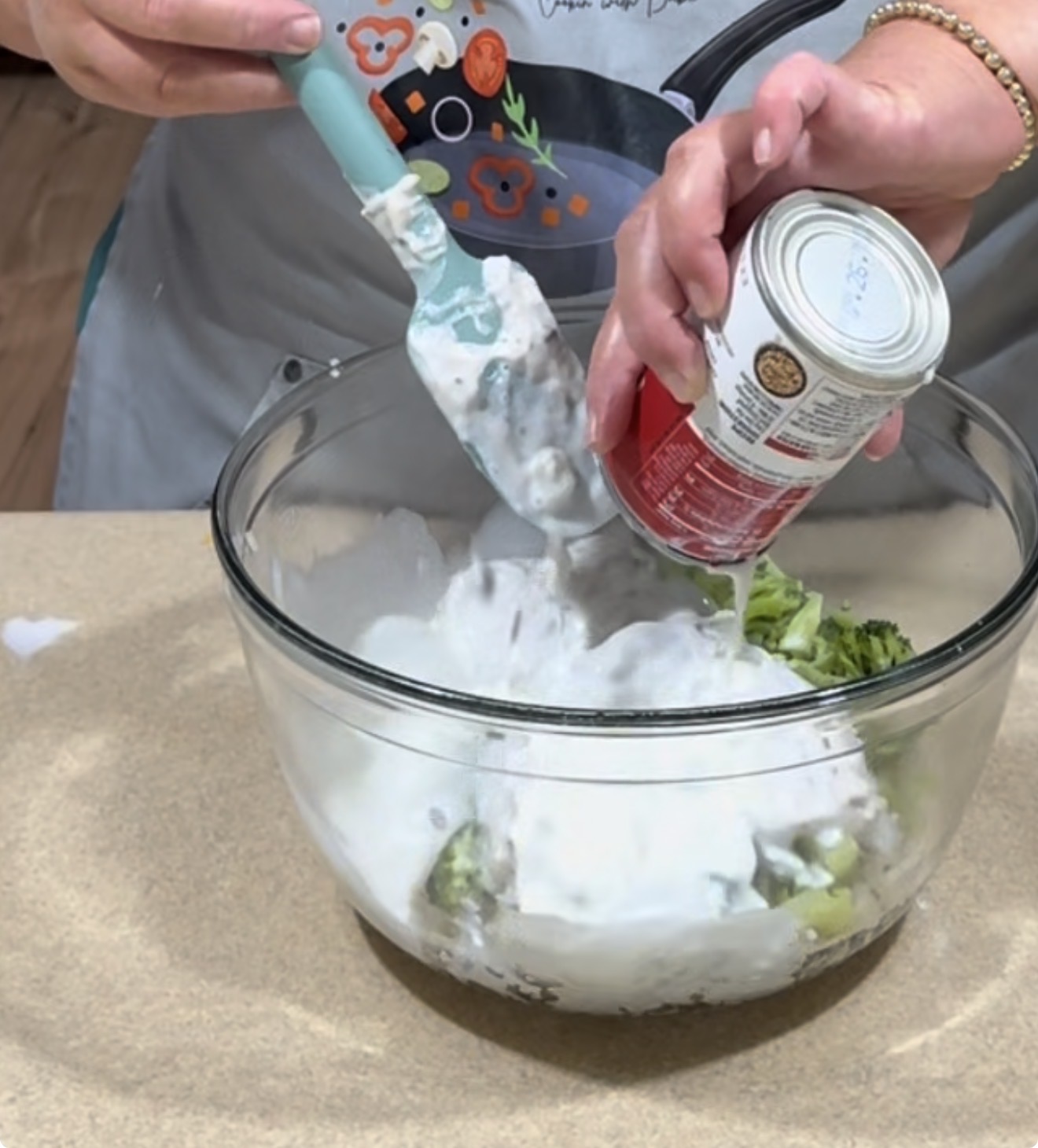 A person pours creamy soup from a can into a glass bowl of broccoli, using a blue spatula to help empty the can. The bowl sits on a kitchen counter.