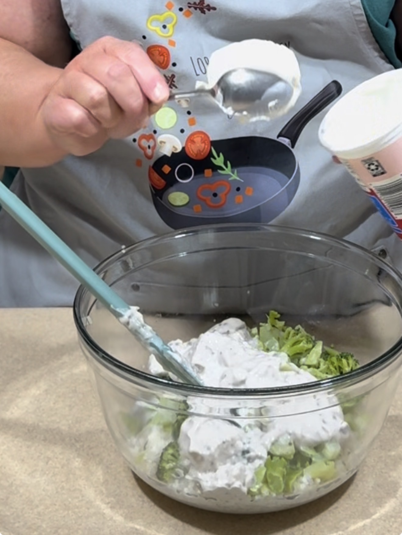 A person wearing a colorful apron is spooning a creamy mixture from a container into a glass bowl filled with chopped green vegetables, preparing a recipe. A silicone spatula rests inside the bowl.