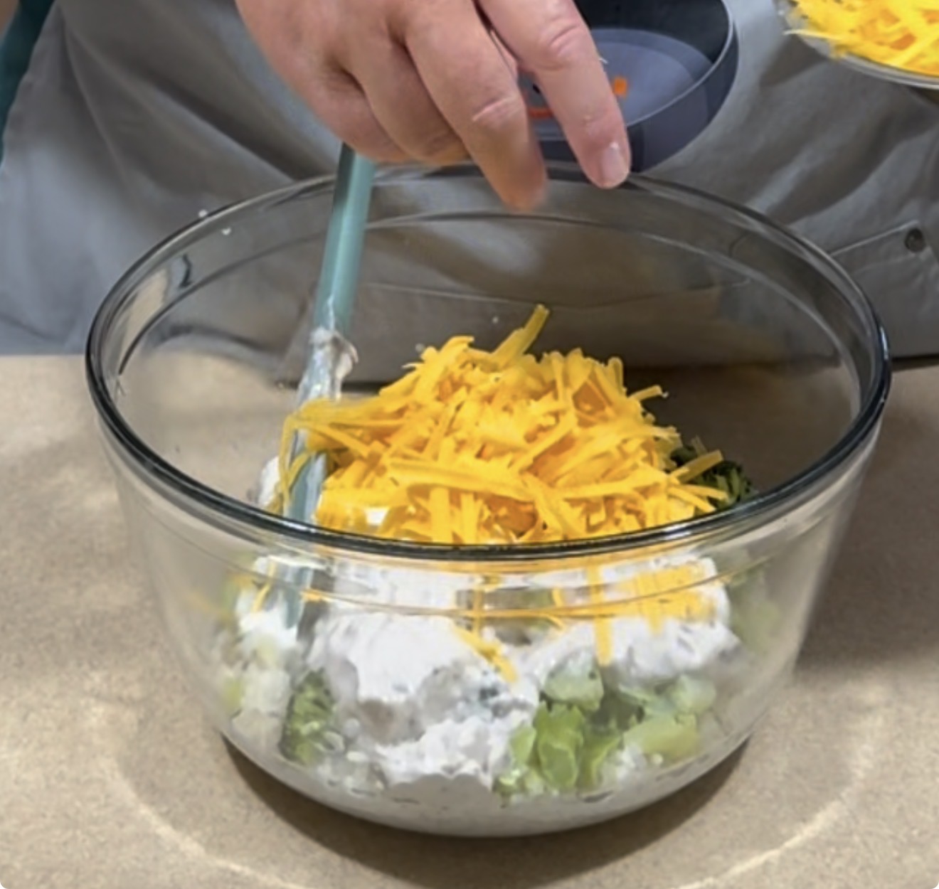 A person is mixing a salad in a glass bowl, containing chopped celery, creamy dressing, and a pile of shredded cheddar cheese on top.