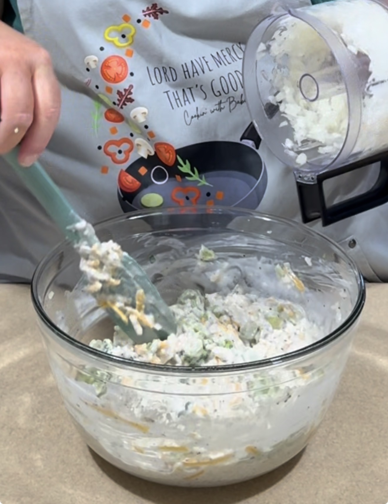 A person wearing a colorful apron stirs a mixture of vegetables, cheese, and creamy sauce in a glass bowl, holding a food processor with chopped onions above the bowl.