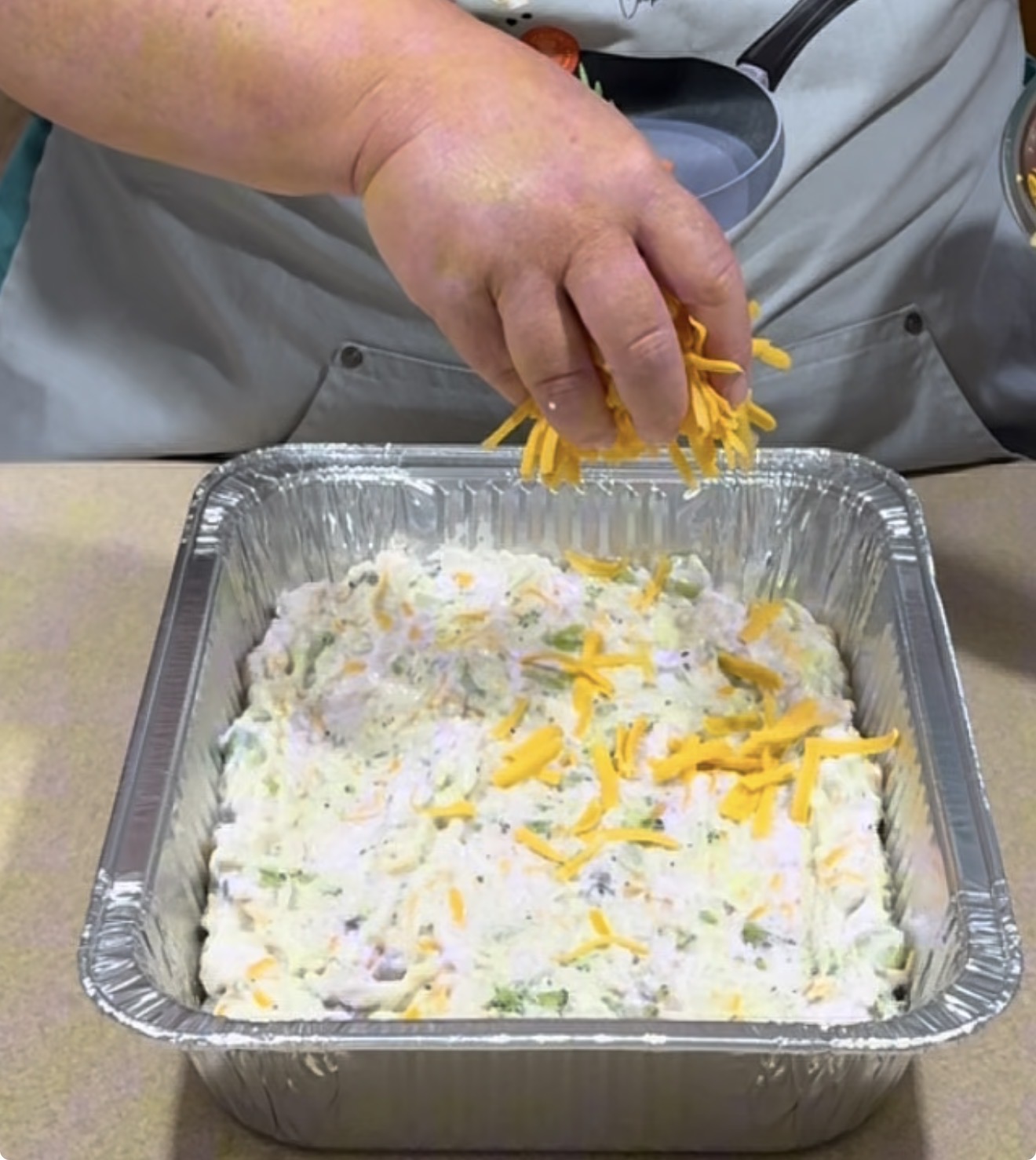 A person sprinkles shredded cheddar cheese over a mixture in a square aluminum baking pan, preparing a dish on a kitchen counter.